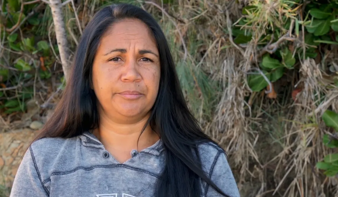 Aboriginal woman with long black hair and wearing a grey top stands in front of foliage growing on a rocky bank.