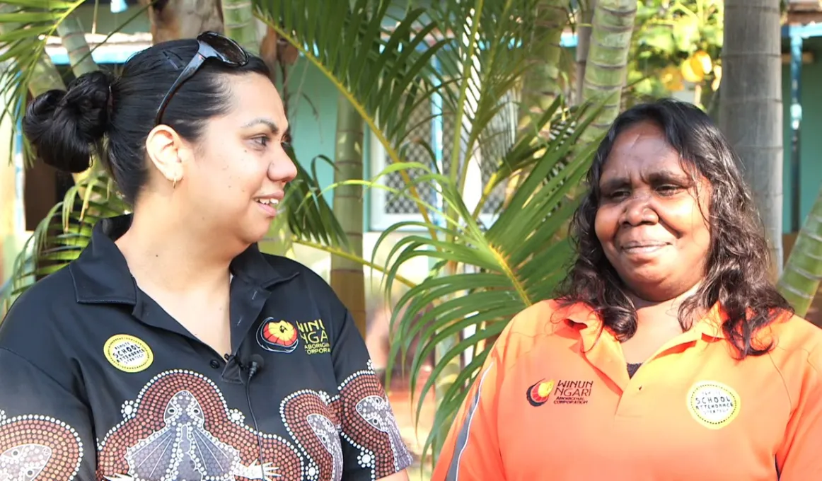 Two Indigenous women standing in front of a palm tree, one wearing a black shirt with Aboriginal design and the other an orange shirt.