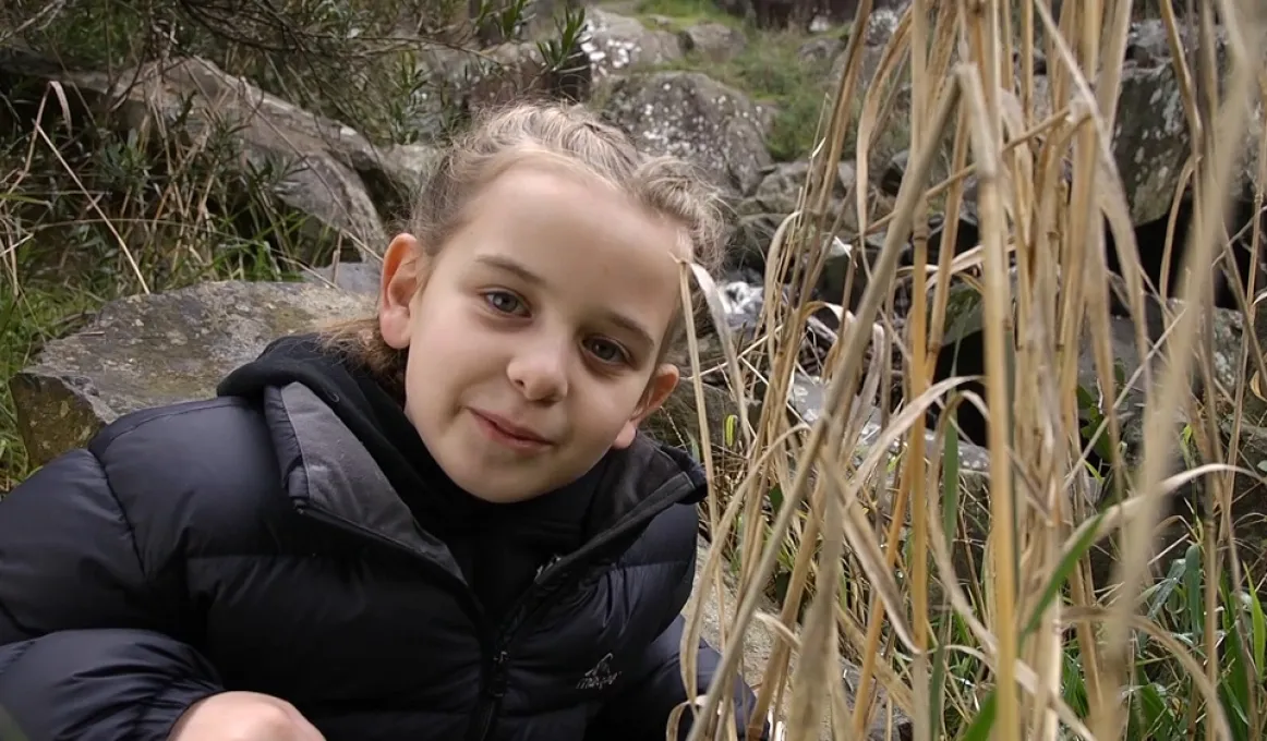 Young girl in a black jacket looks to camera. Beside her are yellow reeds and behind are rocks.