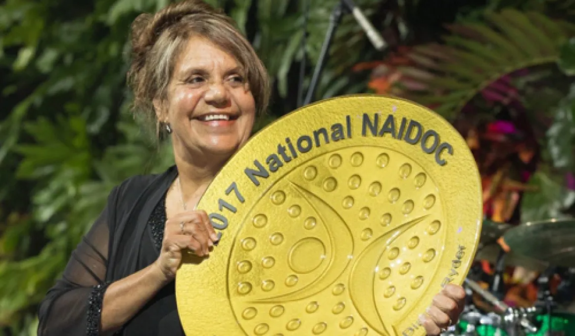 An Aboriginal woman holding a large golden coloured plate with the words “2017 National NAIDOC Lifetime Achievement Award”.