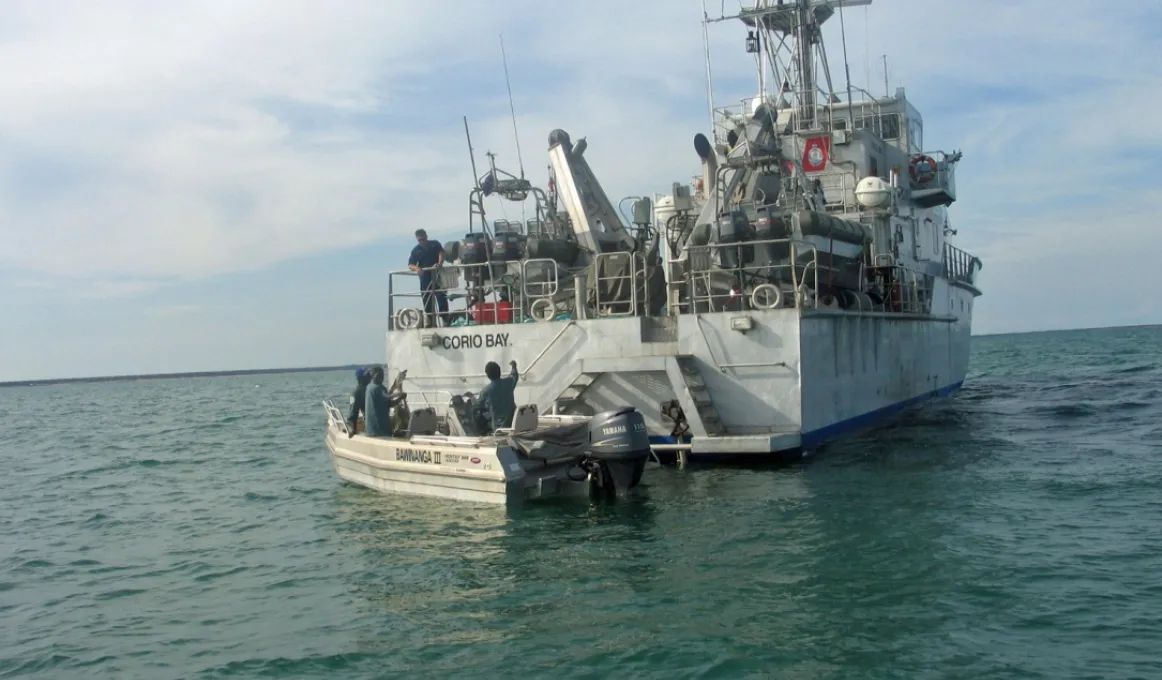 Small motor boat with Djelk rangers in water at back of naval vessel in open sea.