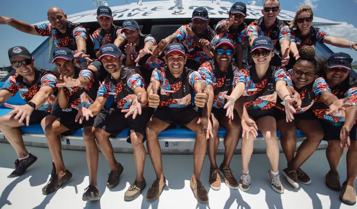 Group of Indigenous adults sitting on the front of a boat and dressed in uniforms displaying an Indigenous design.