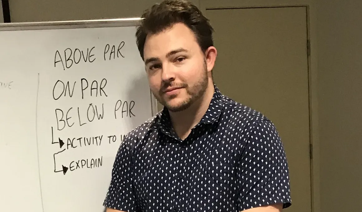 A man in blue shirt with white dots stands next to a white board.