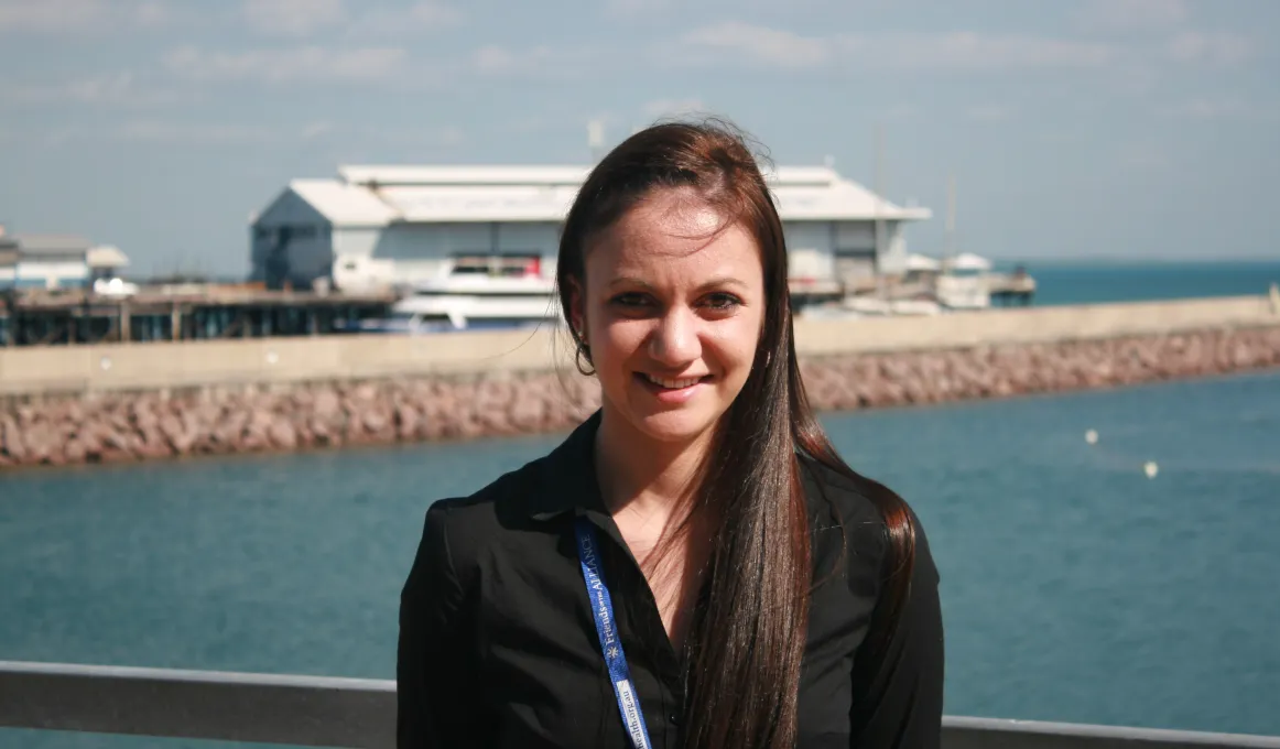 Woman dressed in black shirt stands in foreground with blue water and buildings in the background.