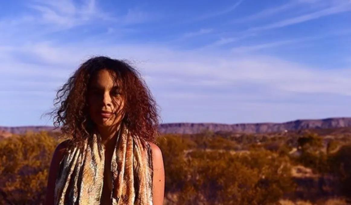 Aboriginal woman with long wavy hair and wearing scarfs around her neck stands in open bushland with a range of hills in the background.