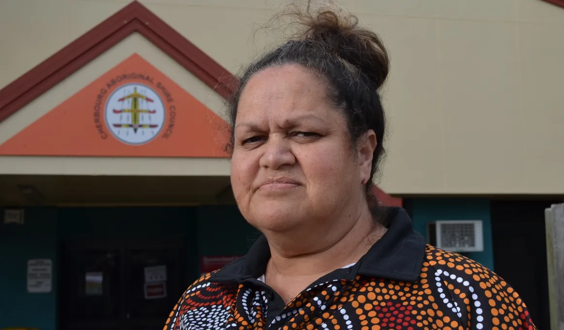 An Aboriginal woman with her hair up and wearing a polo shirt with Indigenous design stands in front of a building.