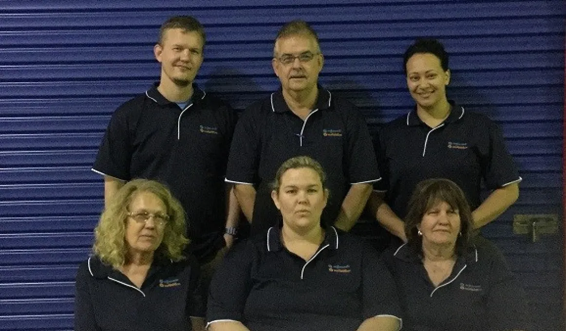 Two rows of people dressed in blue polo neck shirts in front of a blue roller door; three standing in the back row and three are sitting in the front.