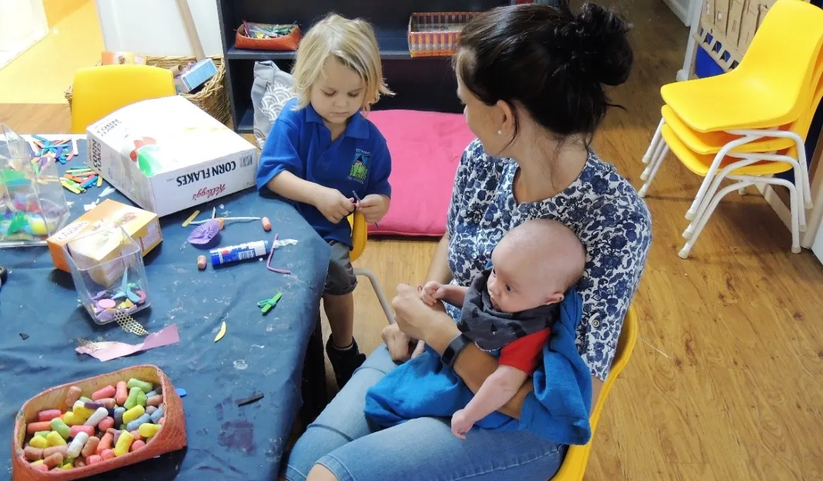 Woman in blue and white blouse sits at small table with craft materials. She holds a baby while looking at a young child in blue shirt working with craft material.