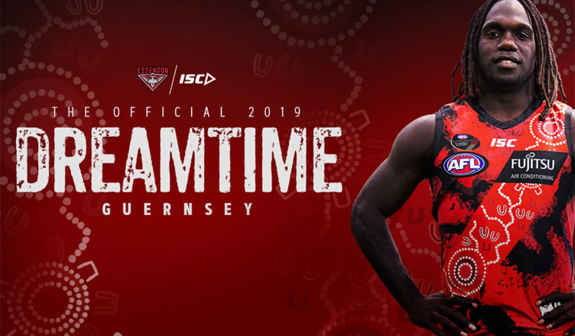 Aboriginal man in red football jumper decorated with traditional designs stands at right. At top left is a logo and the letters: ISC. Below are the words: The official 2019 Dreamtime Guernsey.