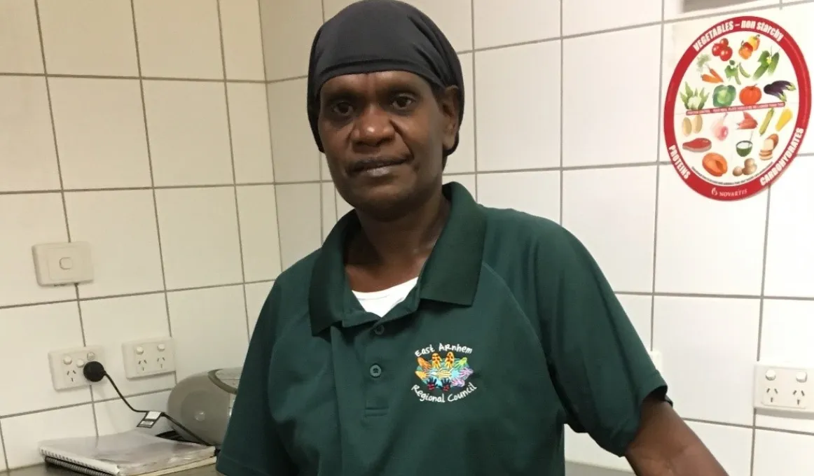 A middle-aged Indigenous woman in green shirt and head covering stands in front of a stainless steel bench with a white tiled wall in the background covered in powerpoints and a poster about fruit and vegetales.