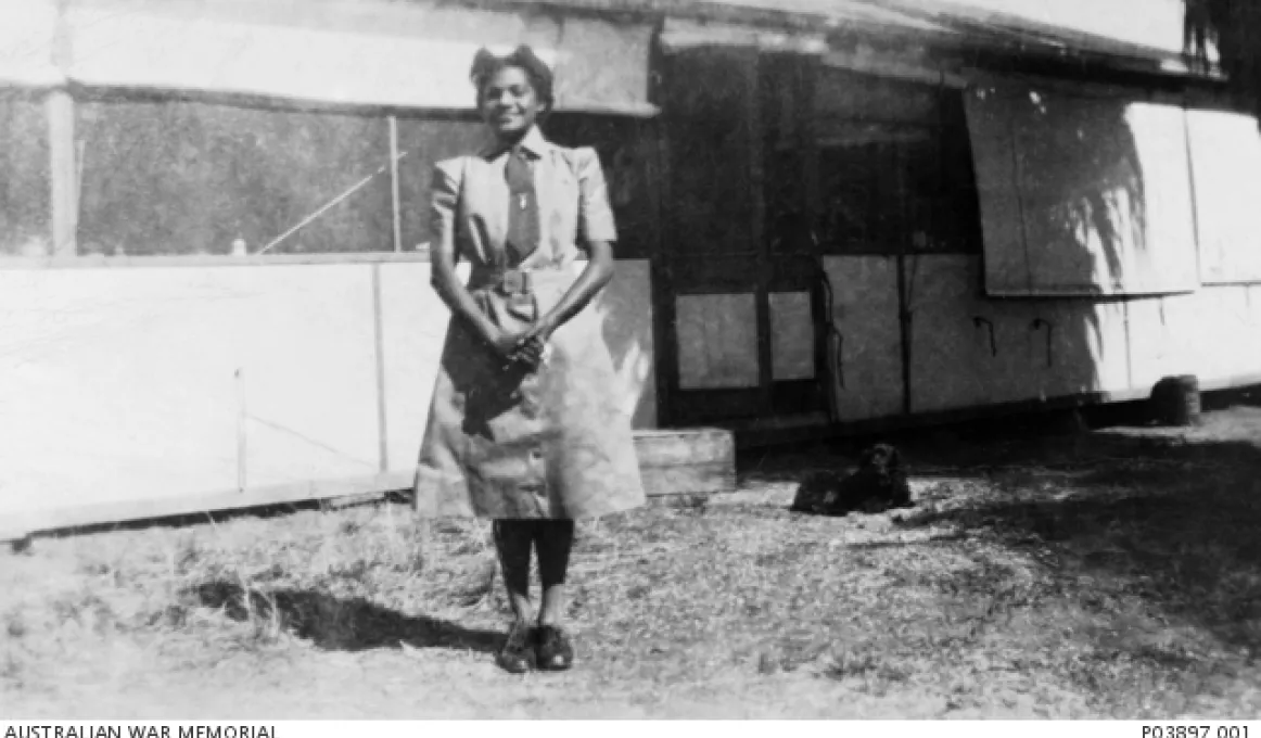 A woman wearing a uniform and hat, stands on grass in front of a building.