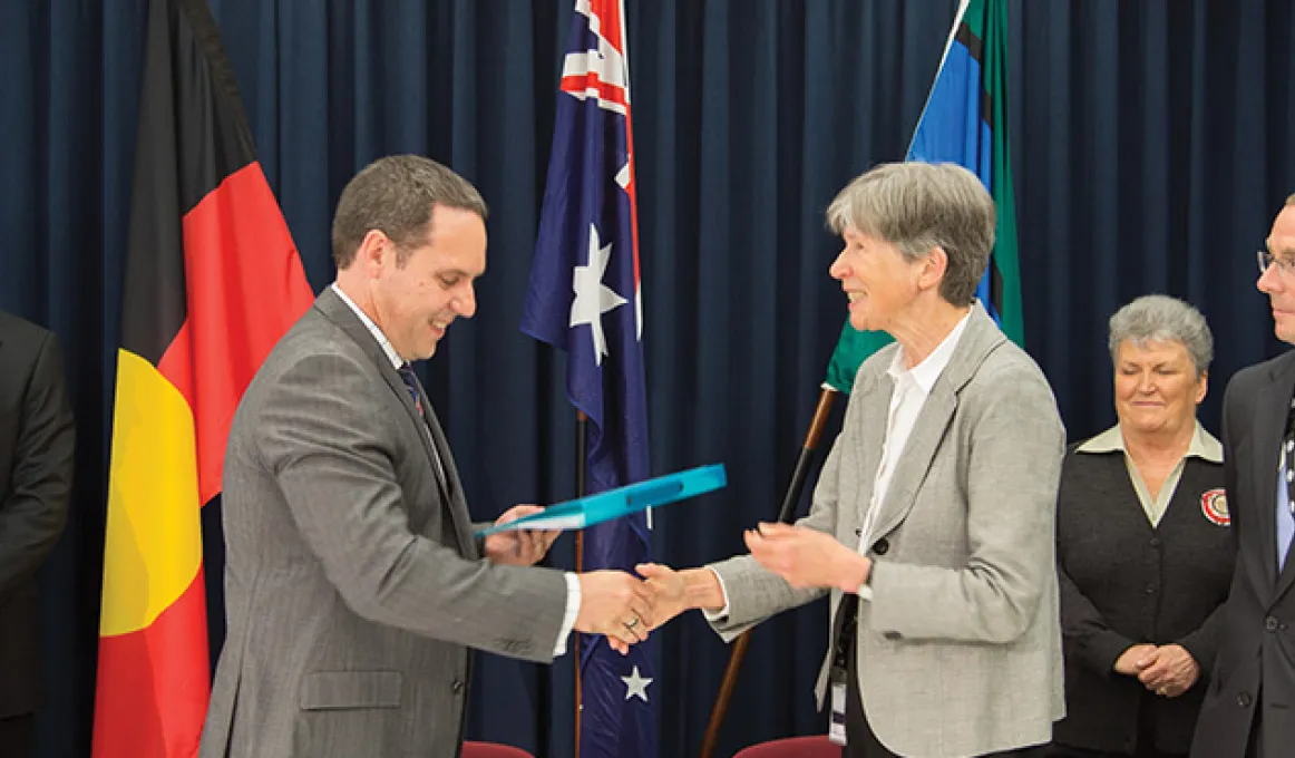 Group of people standing in front of Aboriginal, Australian and Torres Strait Islander flags, watching woman in bone grey coloured jacket shake hands with man in grey jacket.