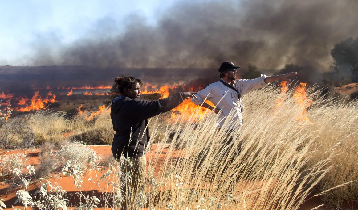 Female ranger and male ranger standing in desert setting with burning grass in the background.