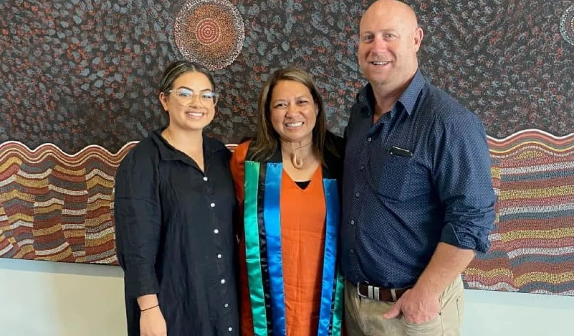 Three people stand shoulder to shoulder. At left is a woman in a dark dress. In the middle is another woman in an orange dress with a green, black and blue sash around her neck and at right is a man in a blue shirt and fawn trousers.