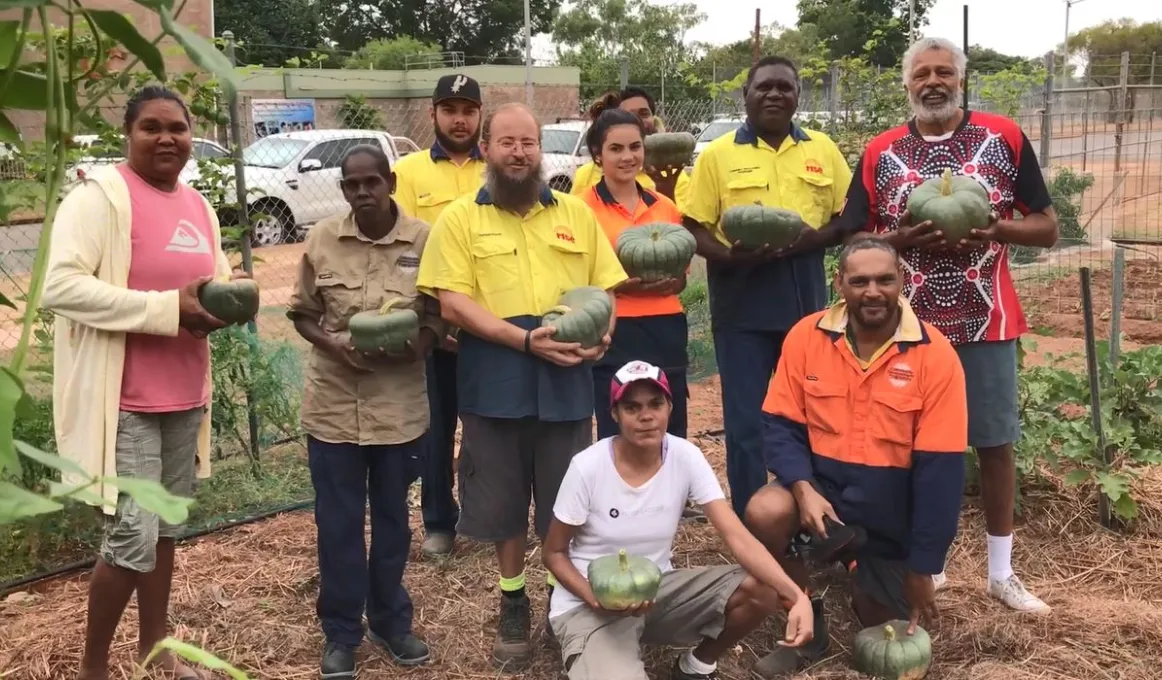 Group of 10 men and women in brightly coloured clothing stand or squat in a vegetable garden, most of them holding a green pumpkin. In the background are cars and buildings and fences.