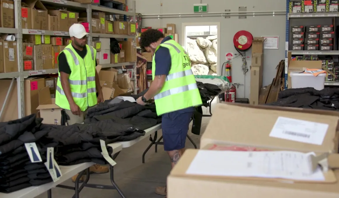 Two men wearing high-visibility vests and blue shorts working in a warehouse filled with boxes. In front of the two men are tables piled with jackets and work clothing.