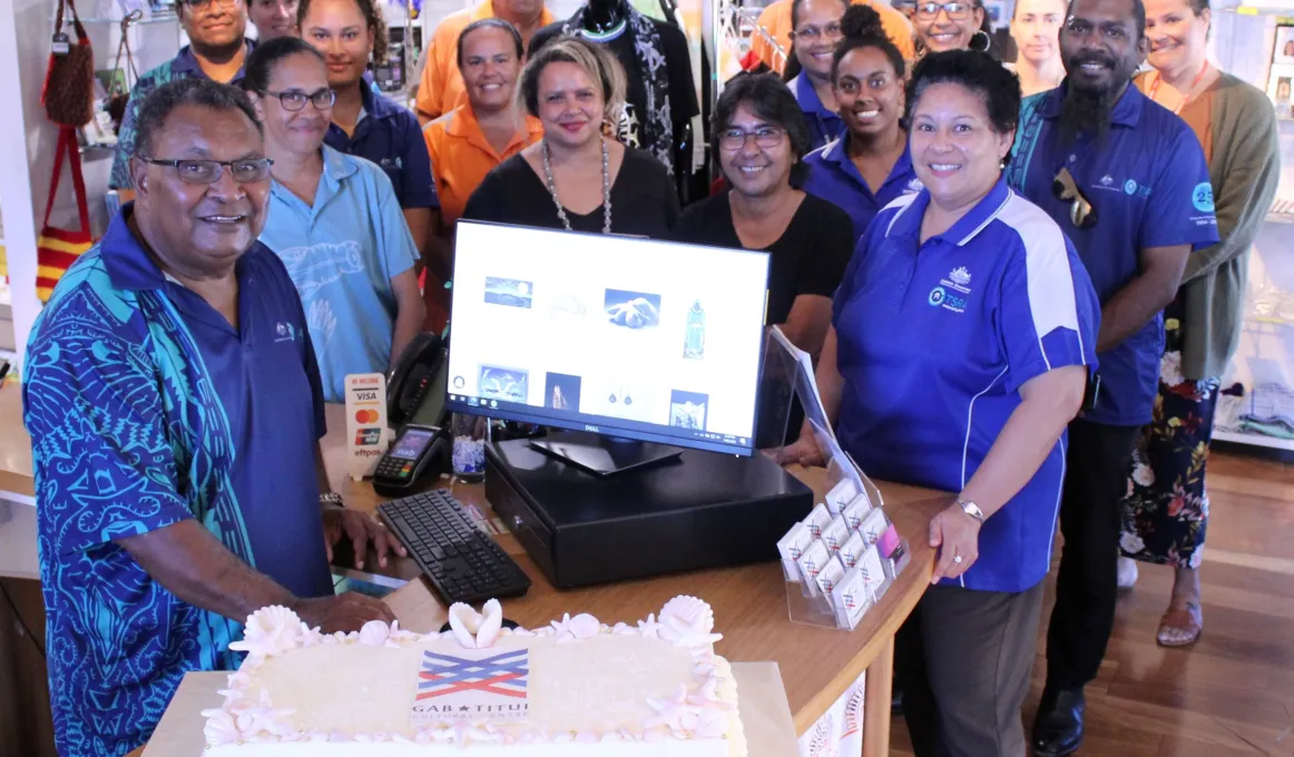 A group of adults in various forms of dress stand behind a large cake. They are in a room with a board floor and on the wall at the back are items for purchase. At the front and behind the cake is a computer.