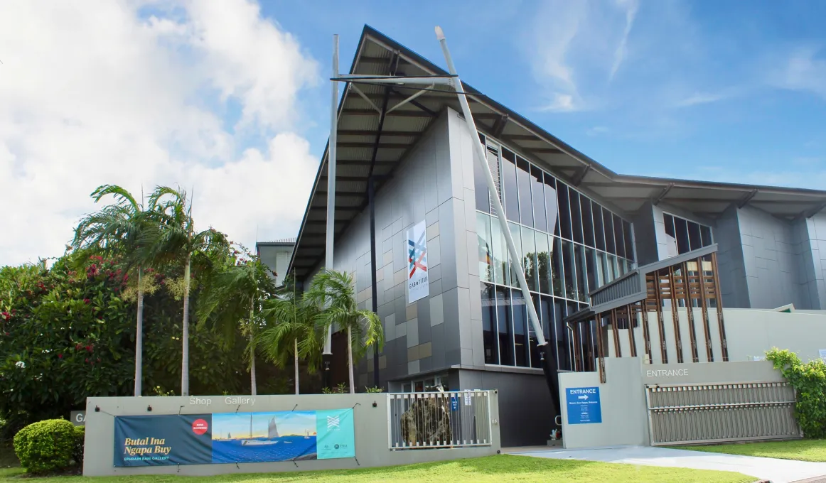 Grey building with many windows. At left are palm trees and others trees and a bush. In front is grass, a walkway and walls.