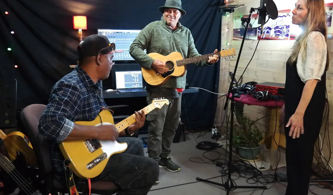 Three people in a small space, wearing casual clothing, are making music. Two men play guitars while a woman stands at a microphone. In the background is recording equipment.