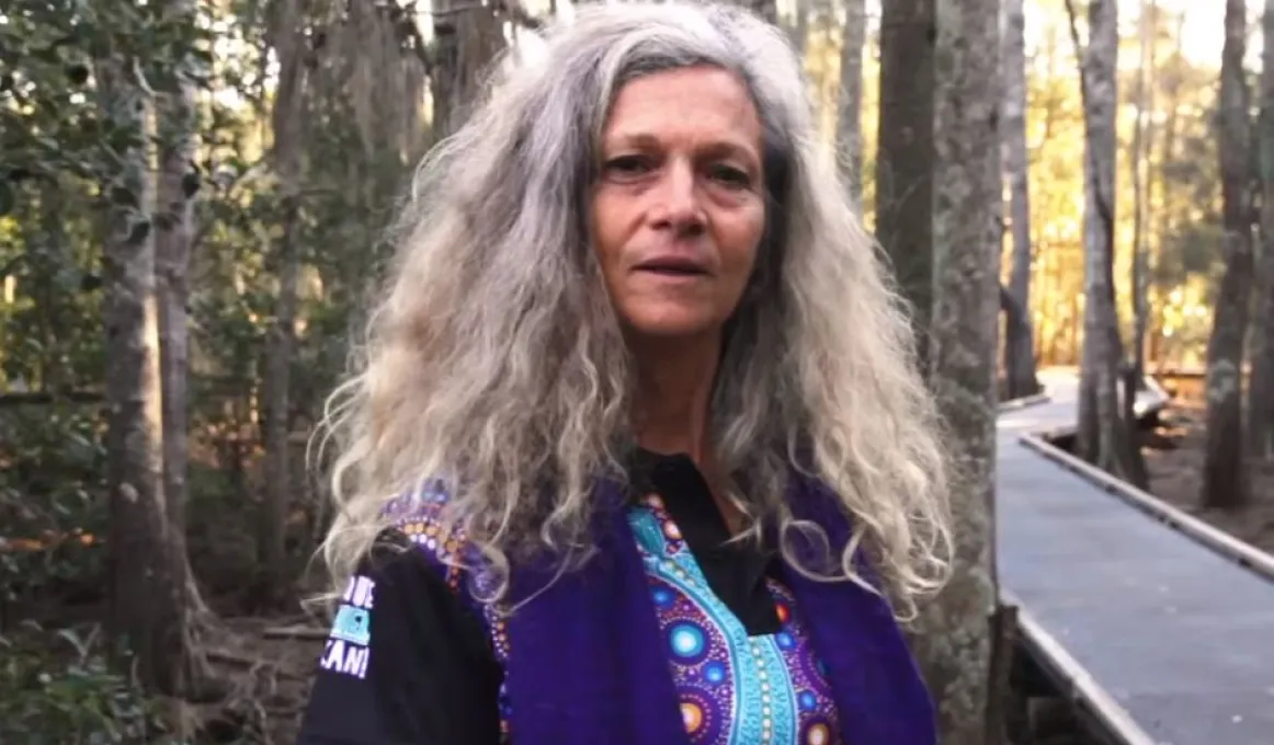 A woman with long greying hair and wearing a purple and blue shirt with Aboriginal designs stands next to a walkway in a forest.