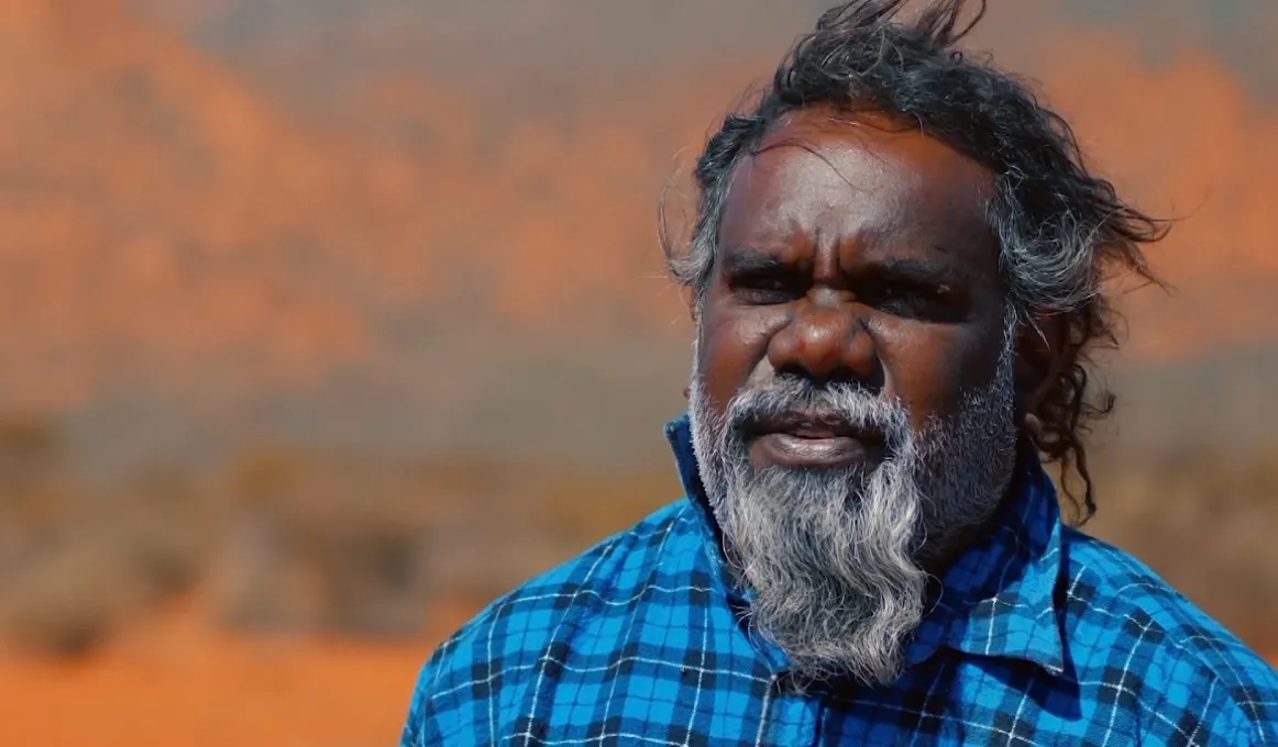 Head and shoulders shot of Aboriginal man with greying beard and wearing a blue checked shirt. In the background is an arid and ochre coloured landscape.