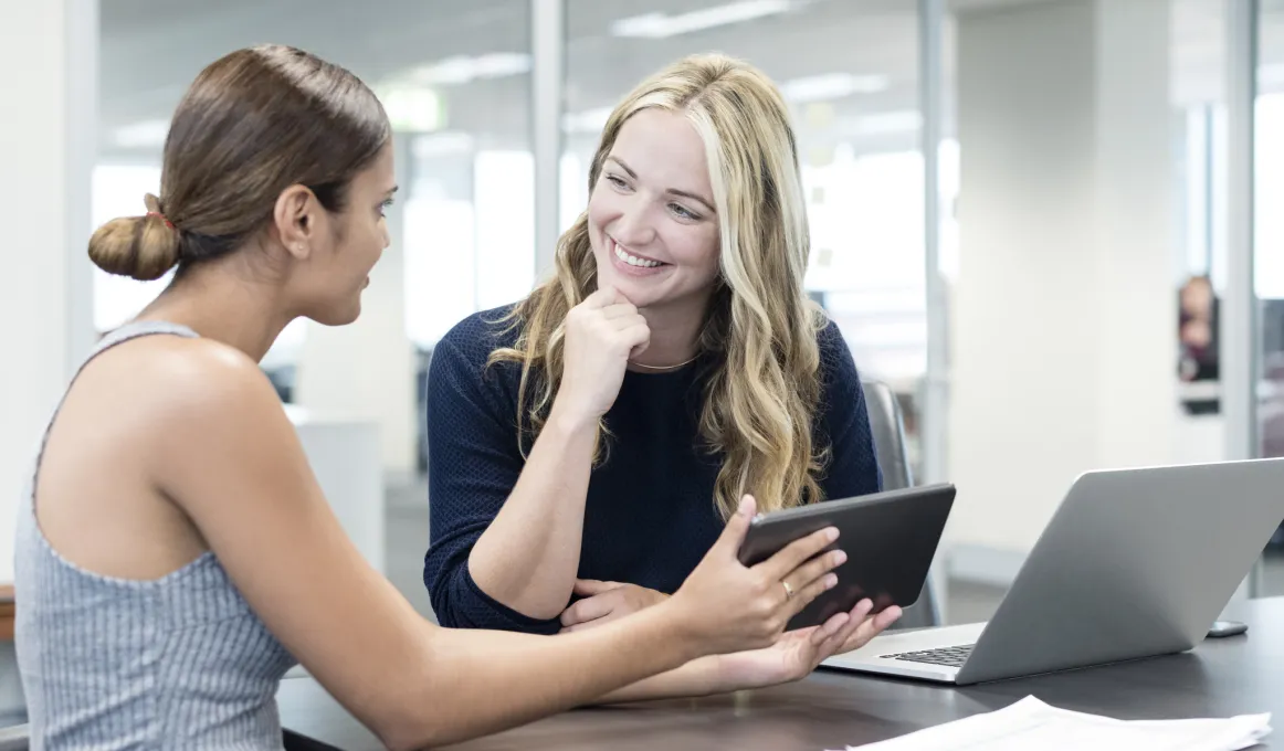 A woman wearing a grey dress is showing a woman in a dark blue shirt something on a digital tablet. They are sitting at a desk with a grey laptop open on the table.