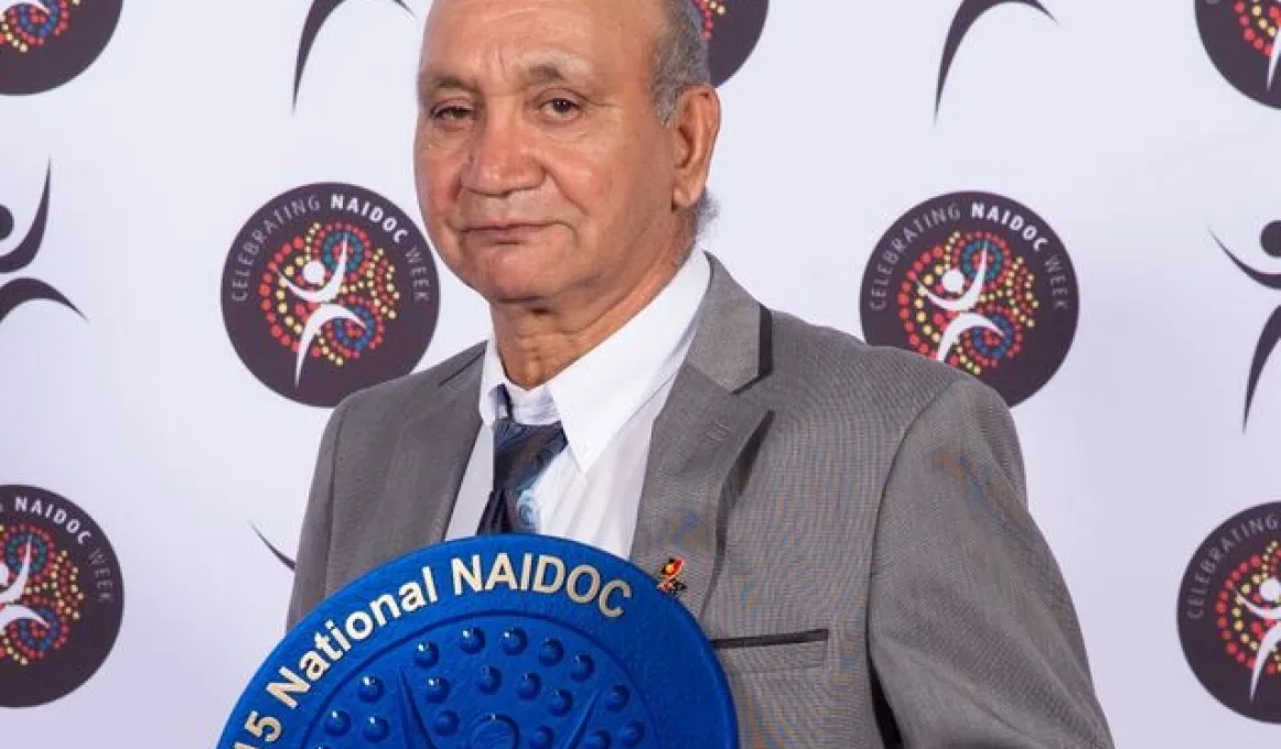 Indigenous man dressed in grey suit holding blue plate displaying the text 2015 National NAIDOC