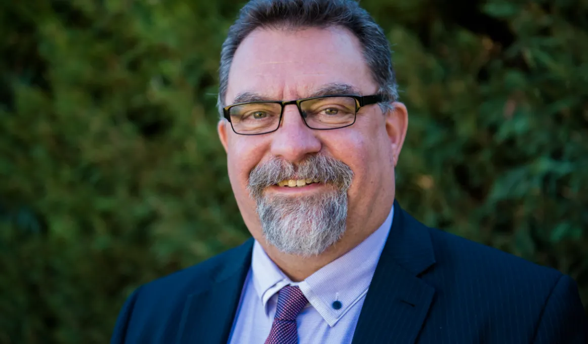 Man with moustache and goatee beard wearing glasses, a blue shirt and dark blue suit stands in front of green foliage.