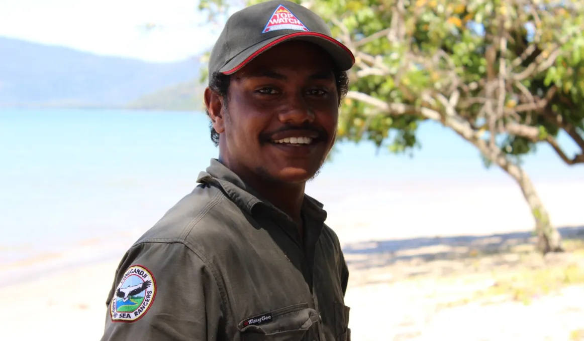 Indigenous young man dressed in ranger clothing standing near tree with body of water and hills in the background.