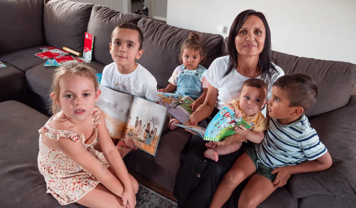 A woman and 5 children sit on a brown couch and look at the camera.