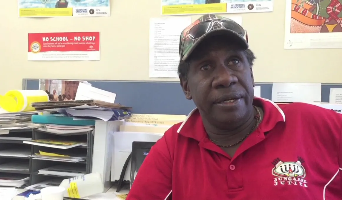 Indigenous man dressed in red shirt and cap seated in an office with posters on the wall behind him.