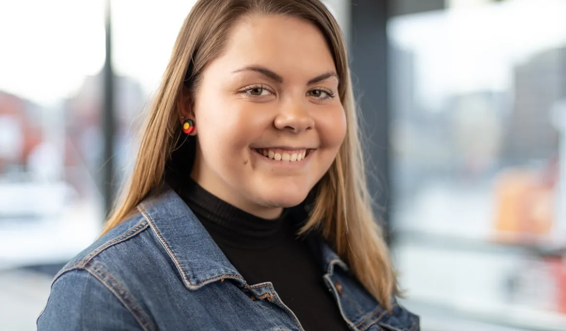 Head and shoulders shot of a young adult woman with long brown hair and wearing a dark top and denim jacket.