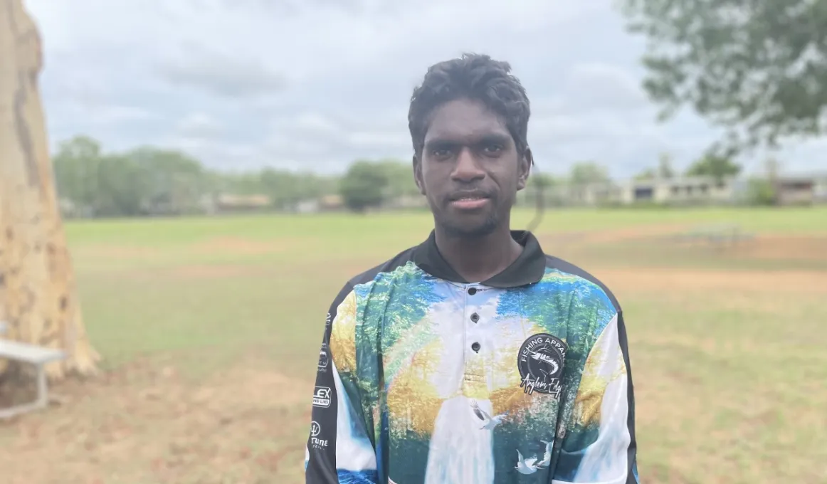 A young Aboriginal man in a patterned, long sleeve polo shirt stands in front of a park or sports oval.