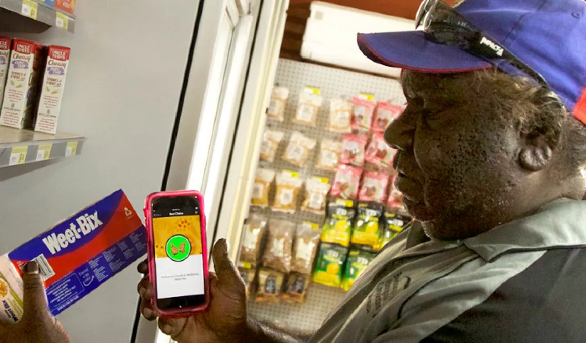 A man wearing a grey shirt and a blue and red cap holds a small packet of Weet-bix in his left hand and a smart phone in his right. In front of him is a shelf of groceries. In the background is a wall with food in plastic bags hanging from it.