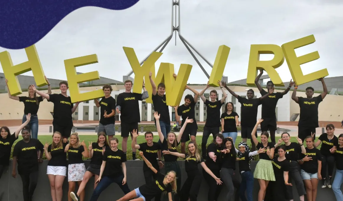 30 young adults are standing in two rows in front of Australian parliament house wearing black t-shirts and smiling. There are 9 young people in the back row holding up the large green letters H E Y W I R E.