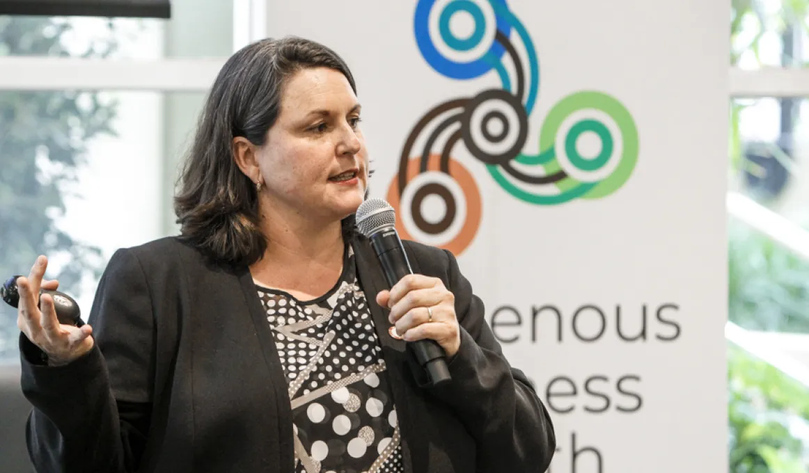 A woman with brown hair and wearing a brown jacket speaks into a microphone. In the background is a large poster in front of a window behind which are trees. The poster has a logo featuring three sets of concentric circles linked by curved lines.