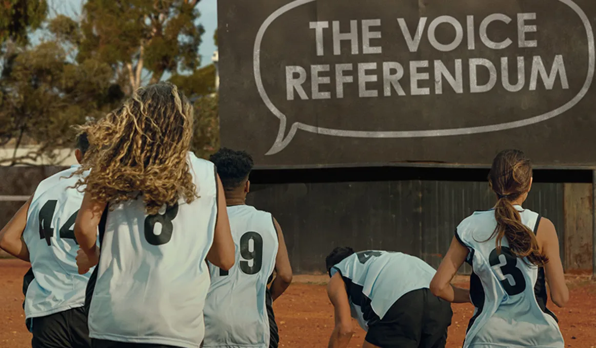 An image of four people wearing sporting jerseys running towards a sign that reads 'The Voice Referendum'.