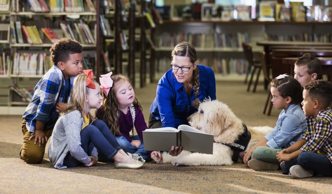 And adult woman sits on the floor holding a book so that children either side of her can see it. In the background are bookshelves.