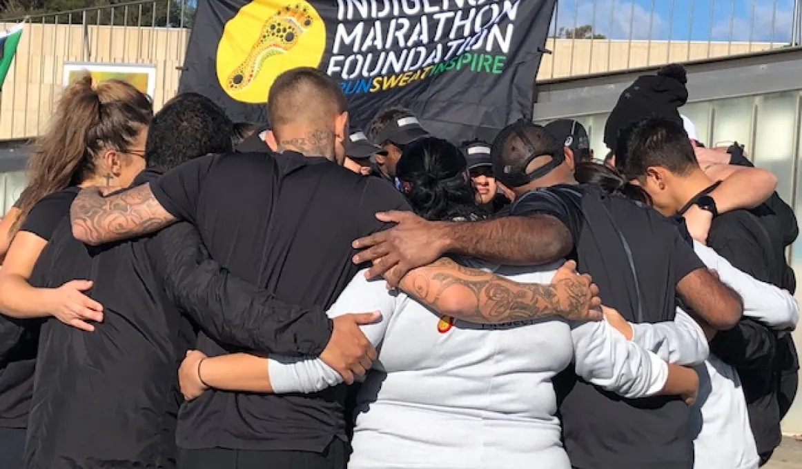 A group of men and women stand in a huddle with their arms wrapped around each other. Behind them is blue sky and a sign moving in the wind. The sign shows the words: Indigenous Marathon Foundation #Run Sweat Inspire