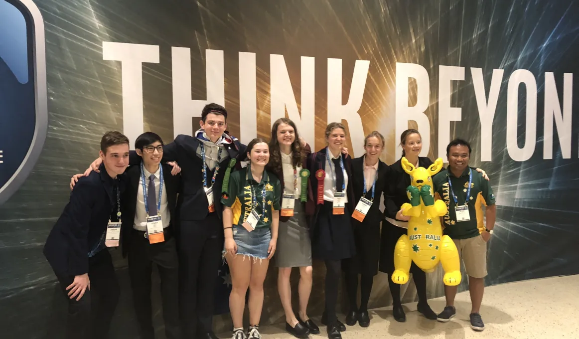 Line of young people arm in arm wearing various clothing styles stand on a hard pale surface and in front of a wall with the words in very large print: Think Beyond.