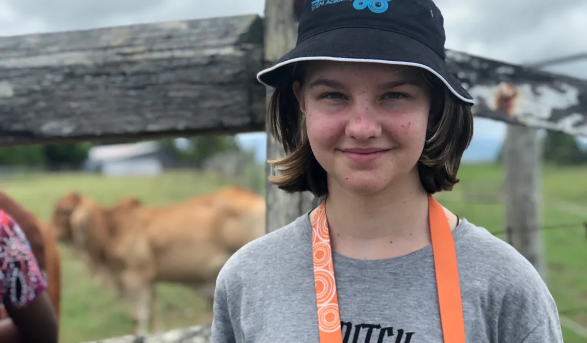 A young girl wearing a grey t-shirt and black hat, looking at the camera. She is standing in front of a paddock with cows.