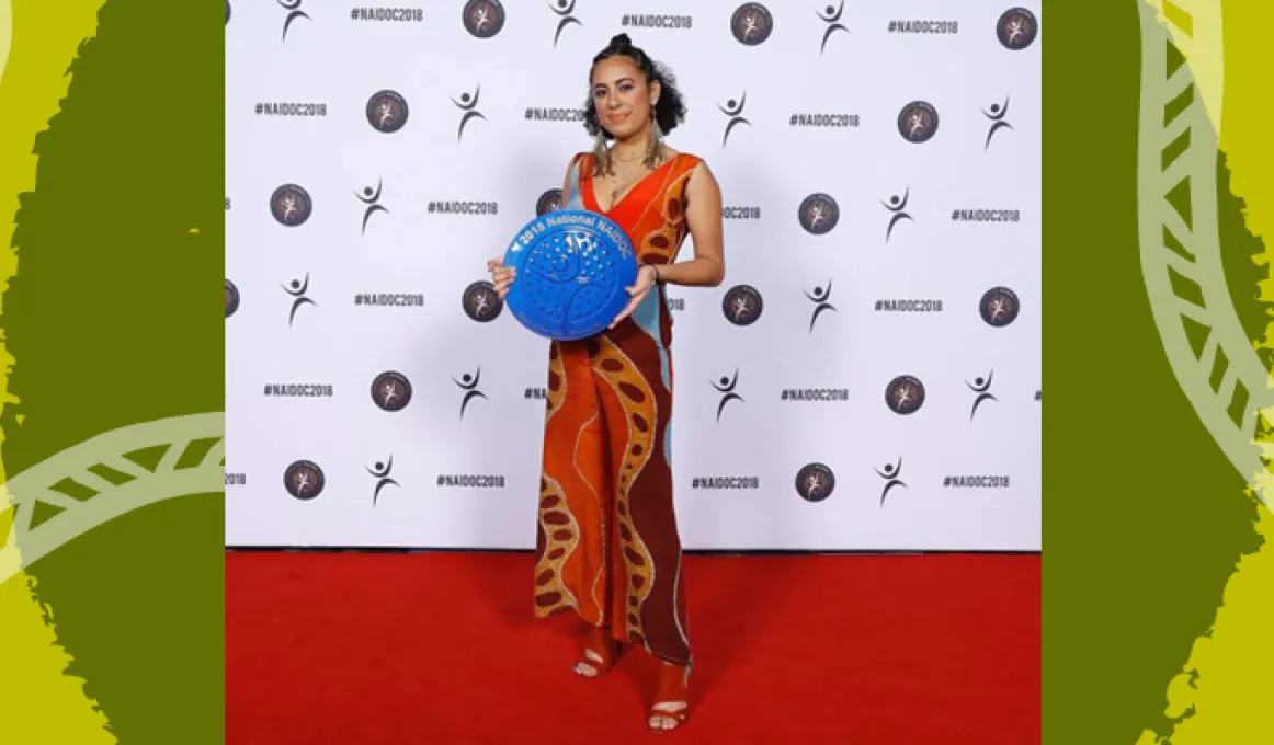 A smiling woman stands on a red carpet, holding a large trophy which is round and vibrant blue with gold writing. It is shaped like a plate. She is standing in front of a wall covered in NAIDOC logos.