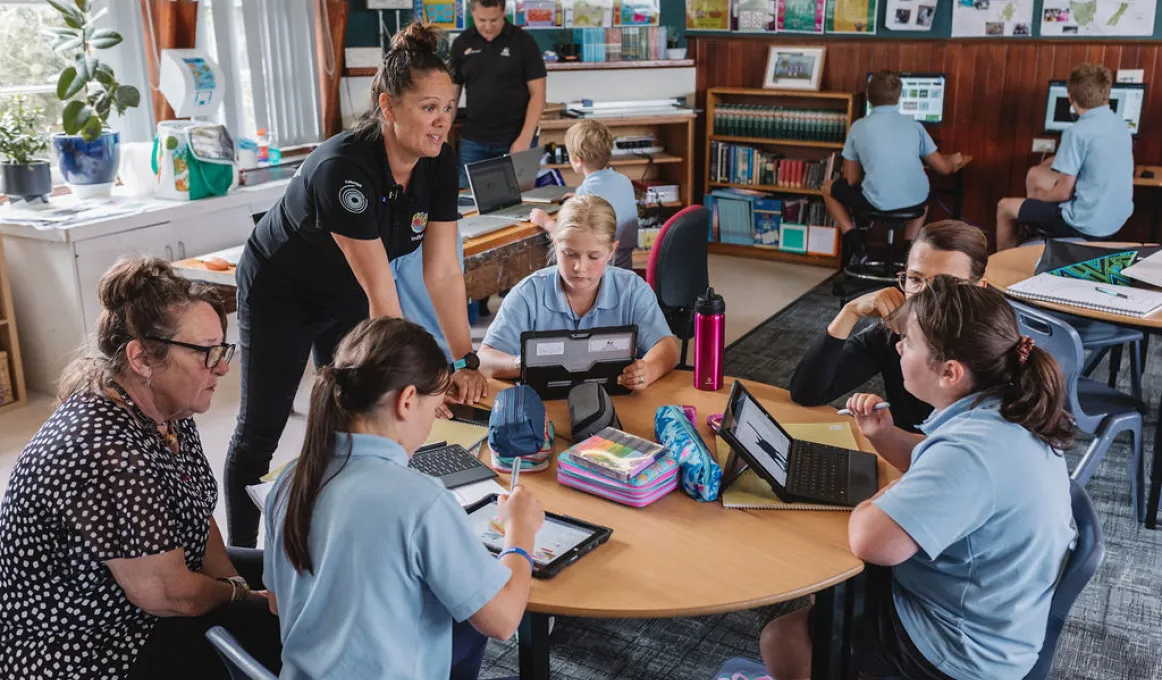 A group of students sit around a table as they work with laptops. A middle-aged woman dressed in black leans on the table while another sits and looks on. In the background are more students, a teacher and classroom paraphernalia.