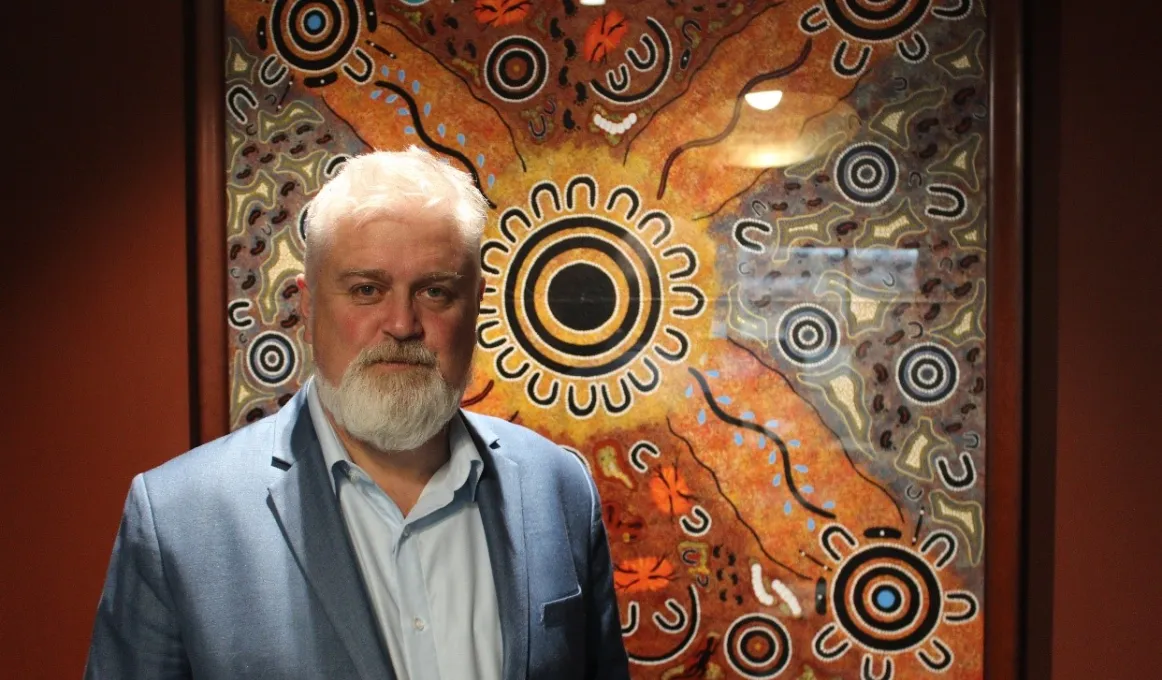 Aboriginal man with white beard and hair, dressed in blue suit and shirt stands in front of artwork of Indigenous design featuring circles, horseshoe shapes and squiggly lines.