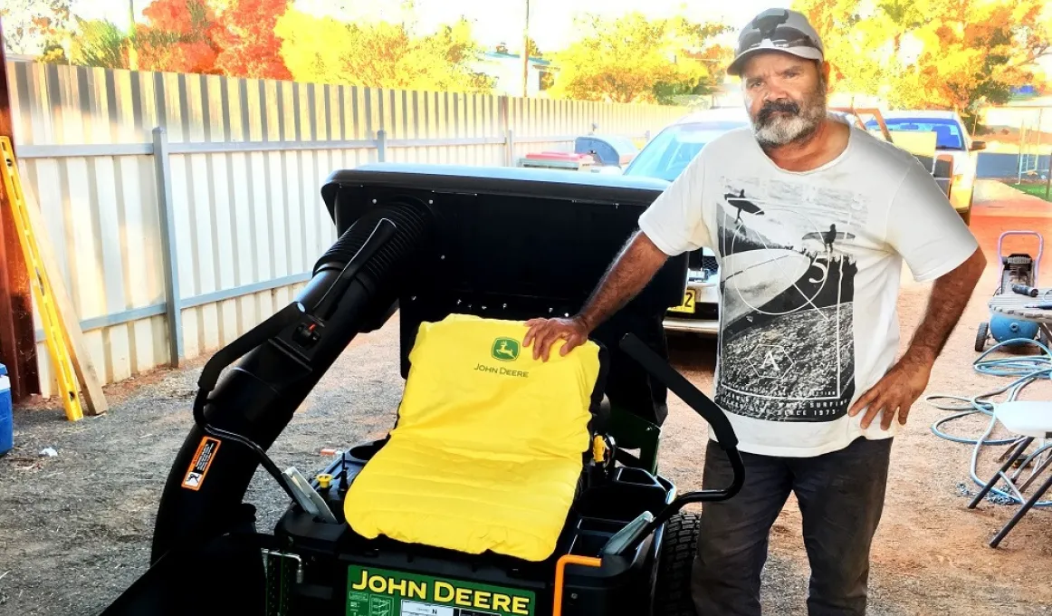 Aboriginal man in white and black t-shirt stands next to a black, yellow and green coloured ride-on lawn mower. In the background is a car, a fence and other equipment.