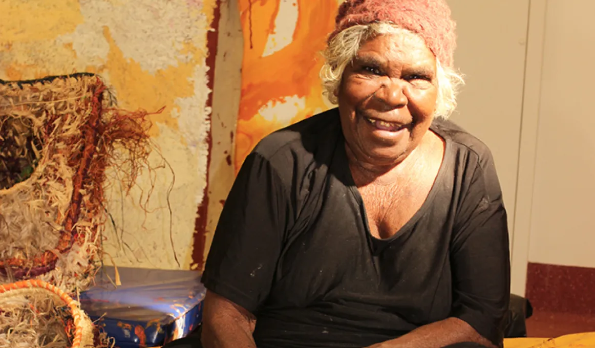 An elderly woman in black dress and reddish beanie is sitting down. In front of her is artwork featuring some birds. In the background is more art and a beige coloured wall.