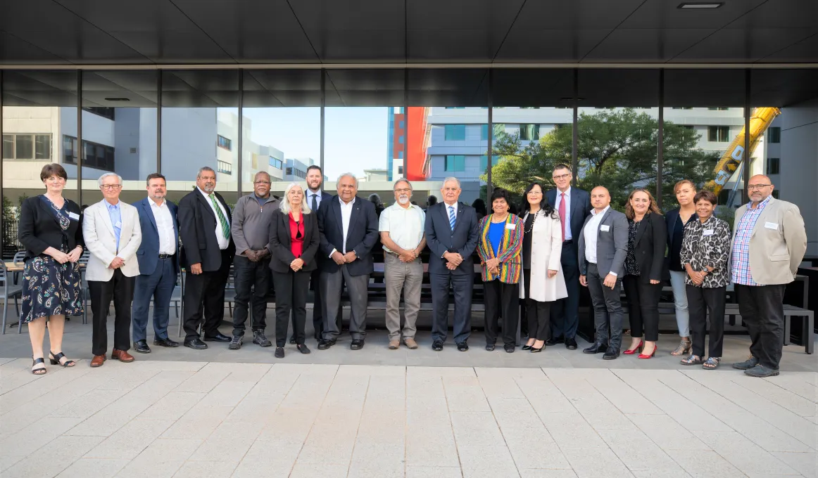 A group of 18 men and women standing in a line in front of a building. The men and women are wearing business clothes and standing with their hands in front of them. In the centre of the group is the Minister for Indigenous Australians.