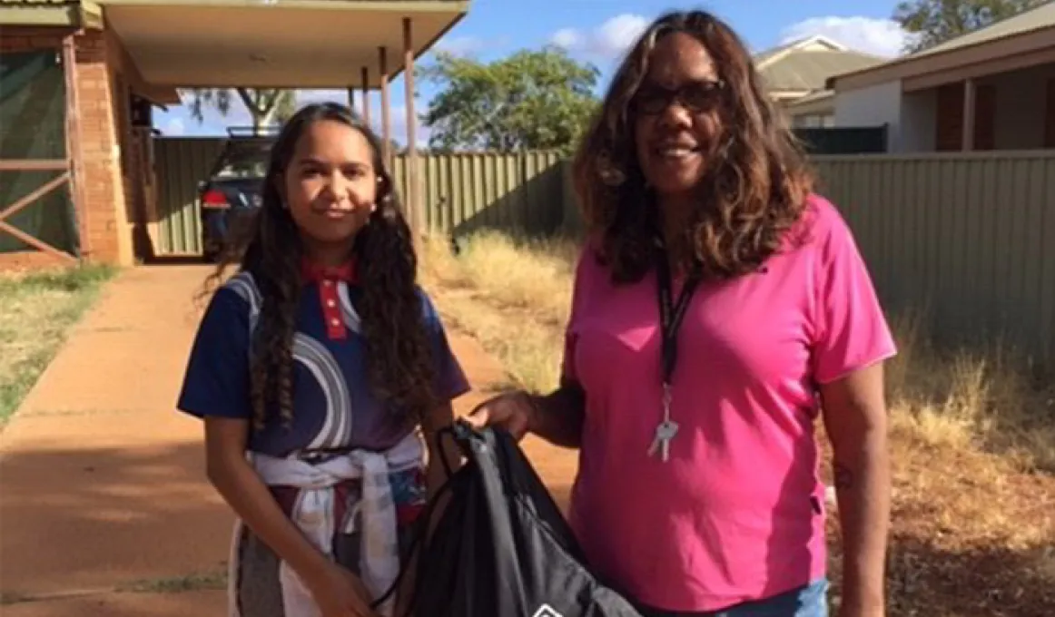 A woman, named Janine Binsiar and a student, Cherice, are standing in a driveway in front of a house and are holding a school and hygiene supply pack.