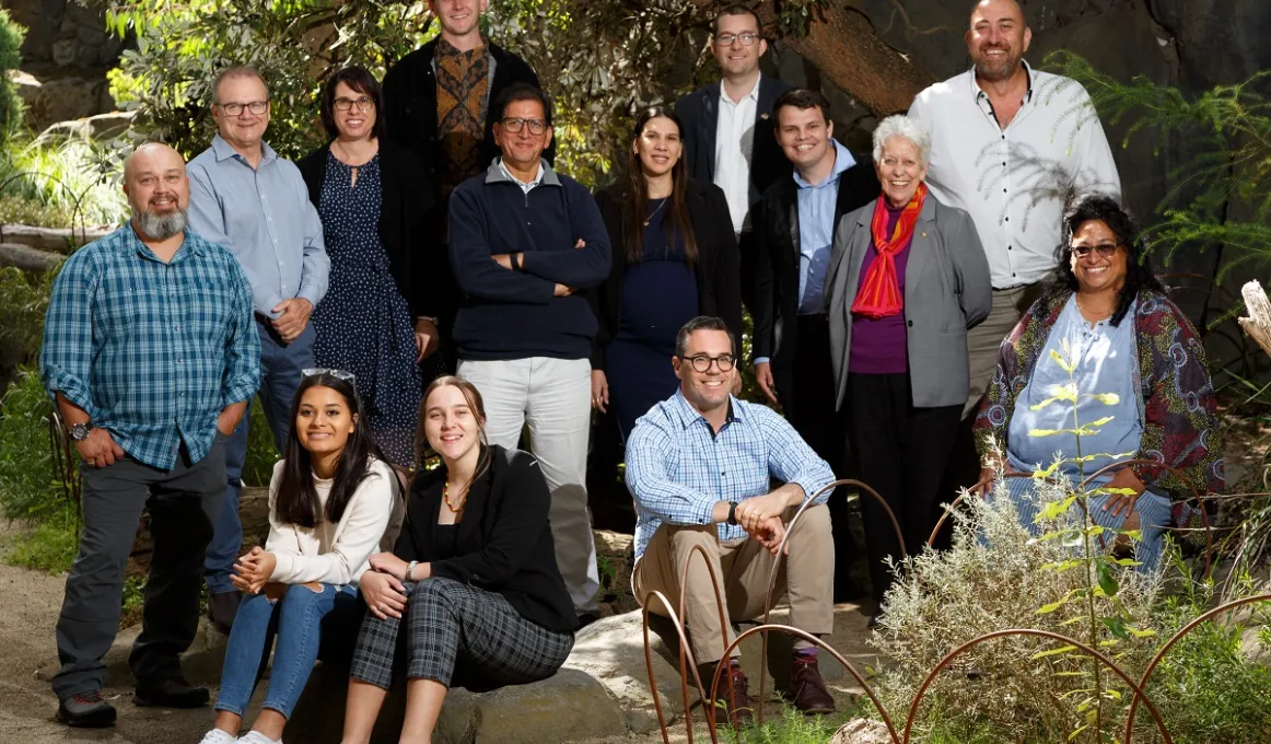 Group of people in various dress types face the camera. Some stand and others sit on rocks. In the background is a tree and other foliage. In the foreground is a fence, rock and plants.