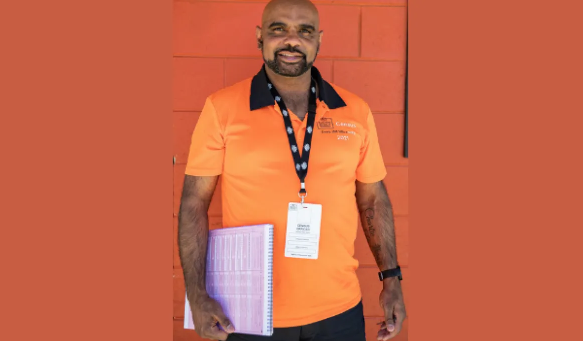 Man standing against brick wall wearing orange 2021 Census shirt and black lanyard holding paperwork.